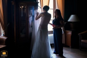   Inside the grand hall at Château du Haget in Gers, the bride focuses intently as she prepares her wedding gown, her concentrated expression reflected softly in the armoire’s glass.